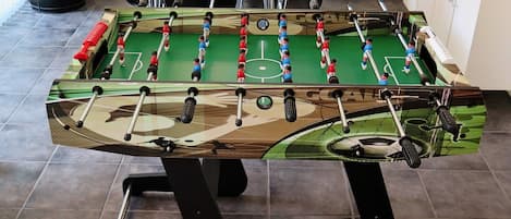 Una mesa de fútbolín con diseño de camuflaje, ubicada en una habitación con suelo de baldosas y una mesa blanca al fondo.