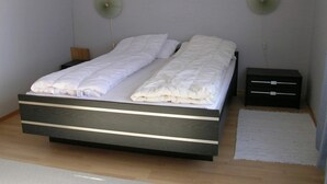 Un llit individual amb estructura de fusta i roba de llit blanca, dues taules de nit i dos ventiladors penjats a la paret.
