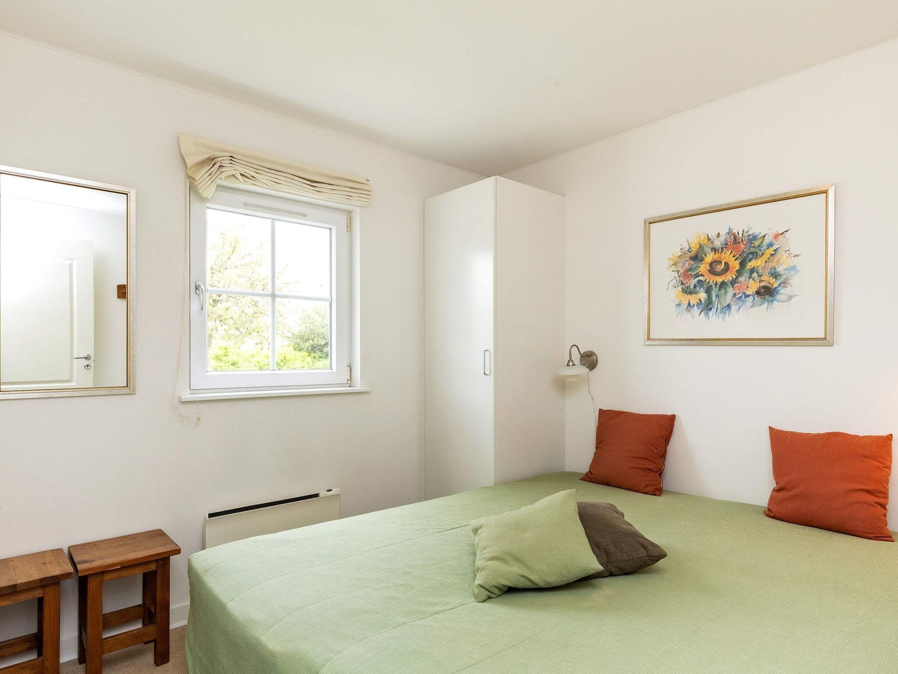 A bedroom with a bed, two pillows, a window, a radiator, a bedside table, and a wall-mounted lamp.