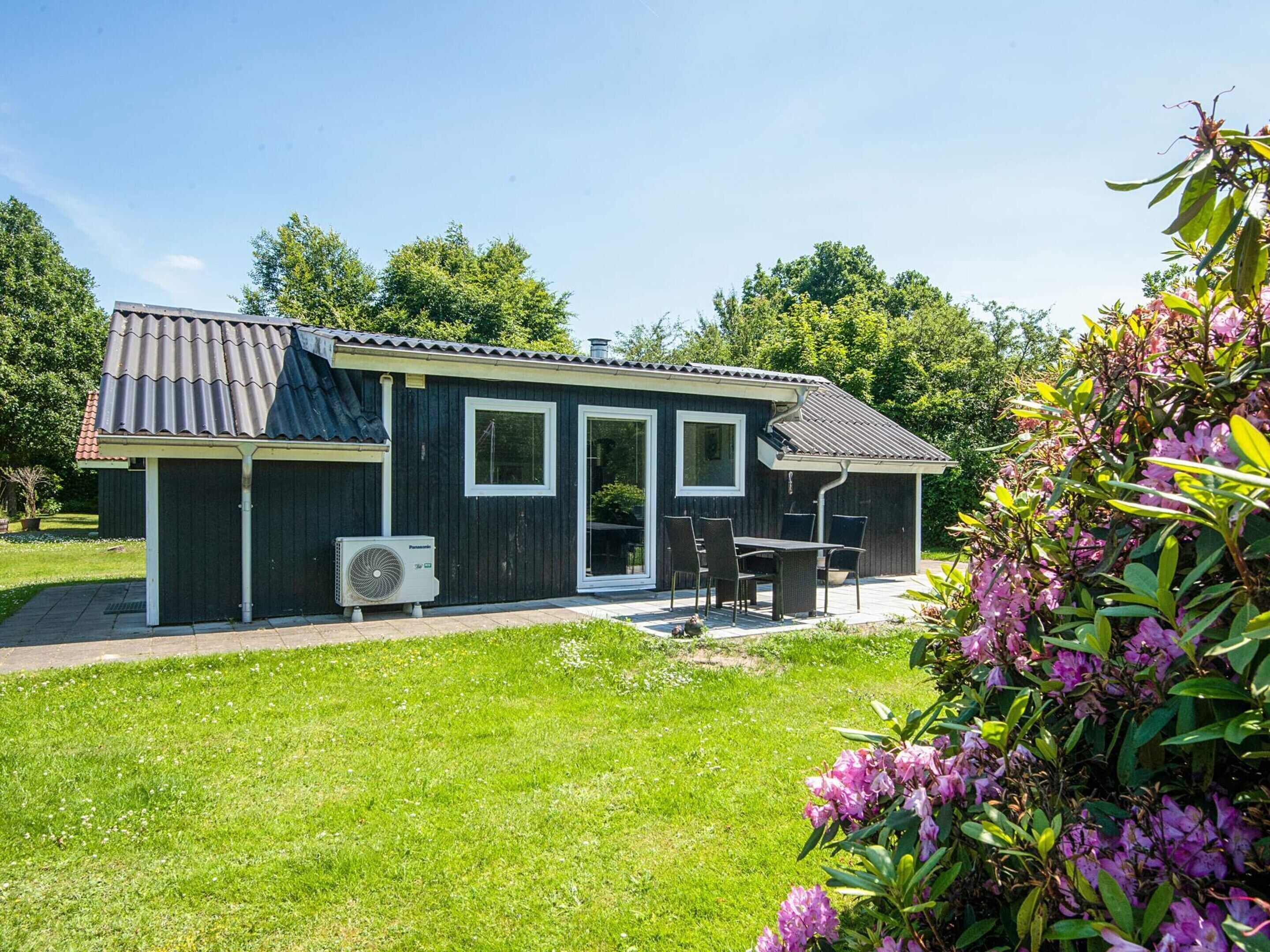 Une petite maison aux couleurs sombres, avec un toit en tuiles, une terrasse avec une table et des chaises, et un jardin planté de fleurs.