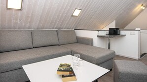 A modern loft bedroom with a sloped ceiling, a grey sofa, a small white table with books and glasses, and a kitchenette area.