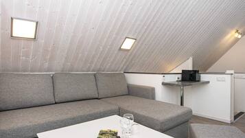 Ein modernes Schlafzimmer im Loft-Stil mit schrägverdeckter Decke, einem grauen Sofa, einem kleinen weißen Tisch mit Büchern und Gläsern sowie einer Küchenzeile.