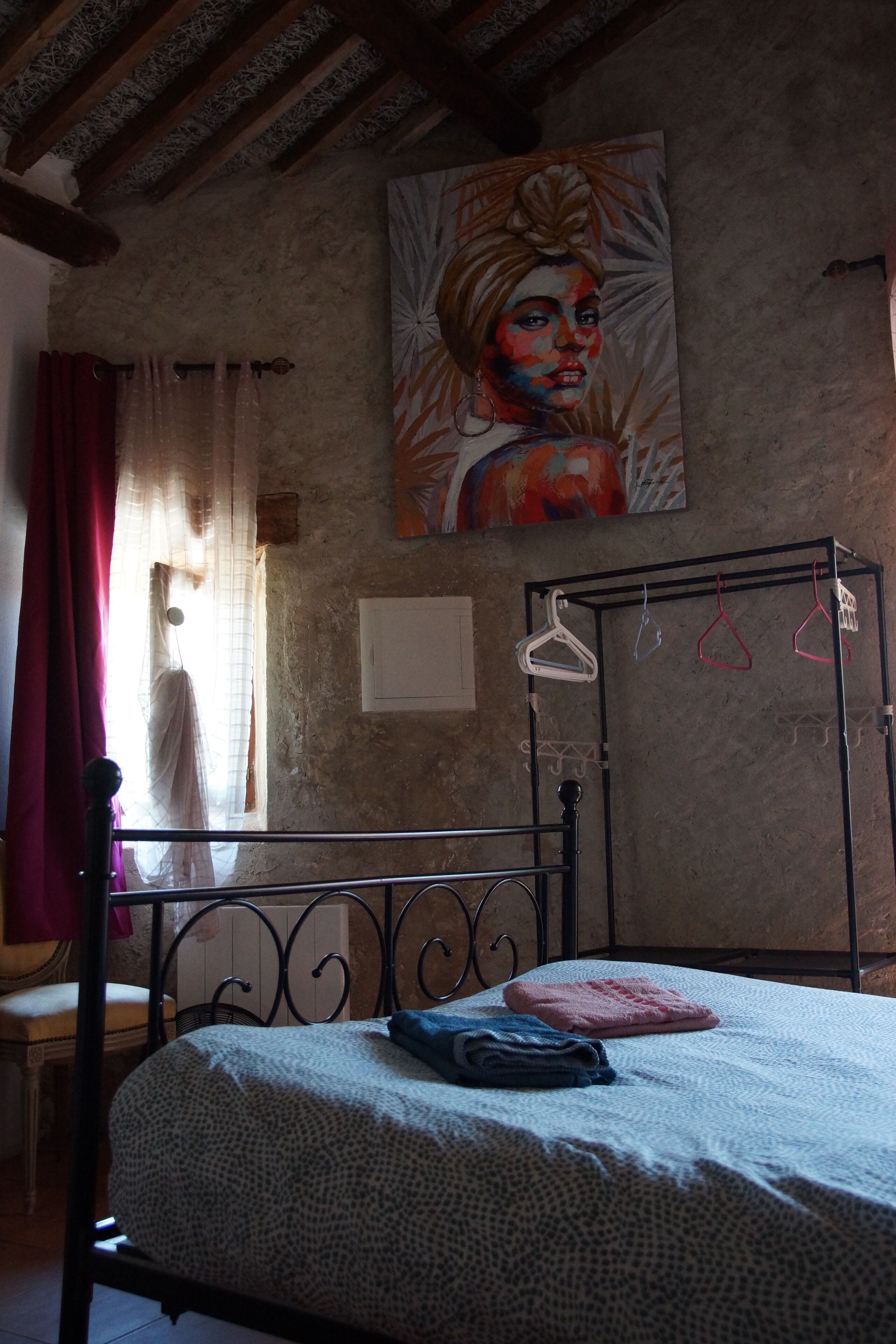 Un dormitorio con cama, armario metálico, un cuadro en la pared y ventana con cortinas.