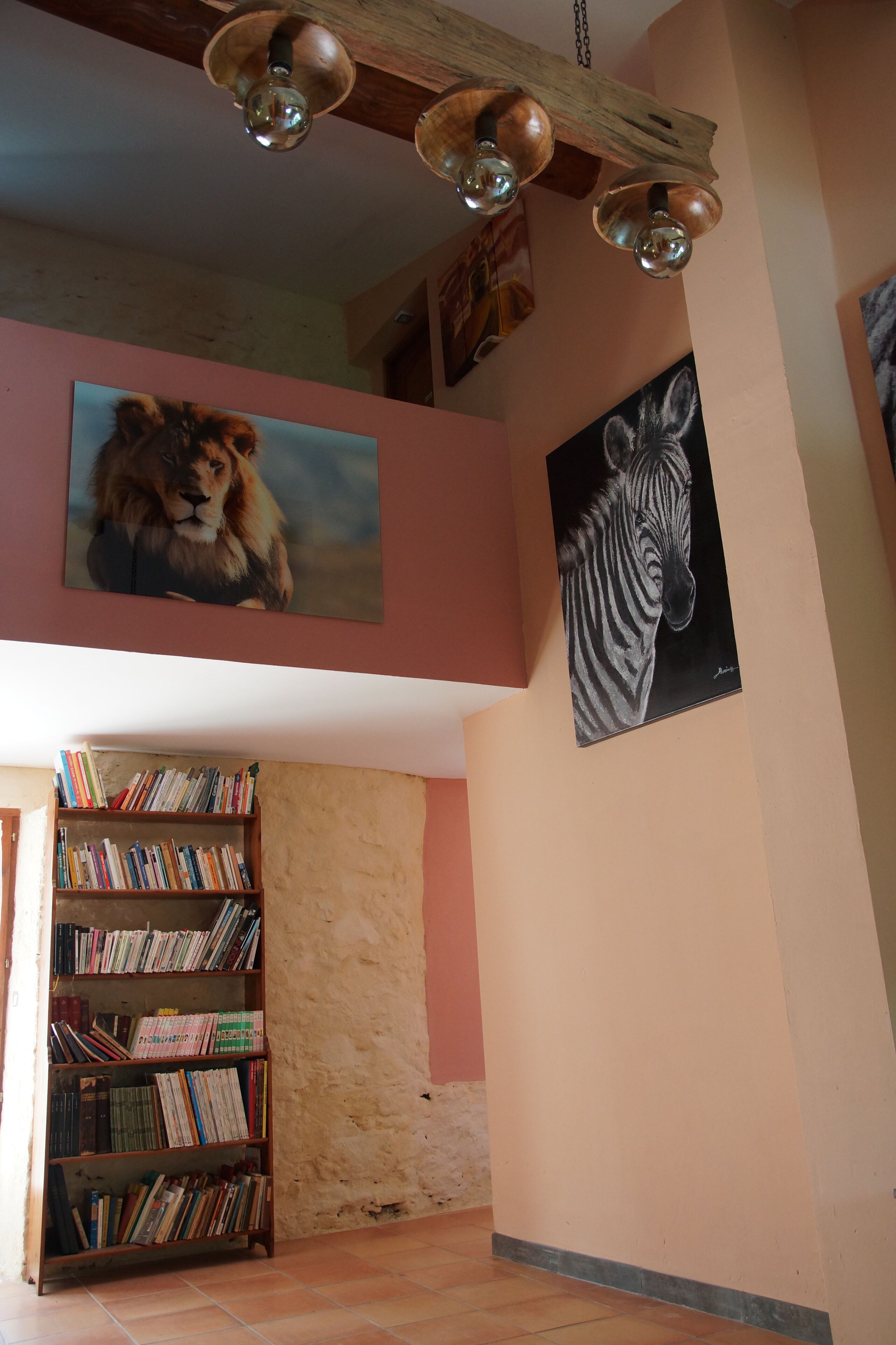 Quarto com teto de madeira, estante repleta de livros e dois quadros emoldurados com animais nas paredes.