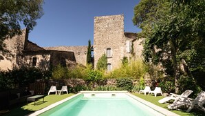 A swimming pool with lounge chairs, surrounded by a stone building with a tower, and lush greenery. - Prieuré d'orniols (Goudargues)