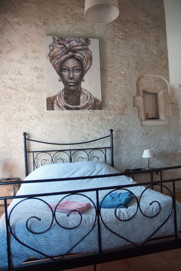 A bedroom with a metal bed frame, a patterned bedspread, a wall-mounted painting, a small table with a lamp, and a stone wall.
