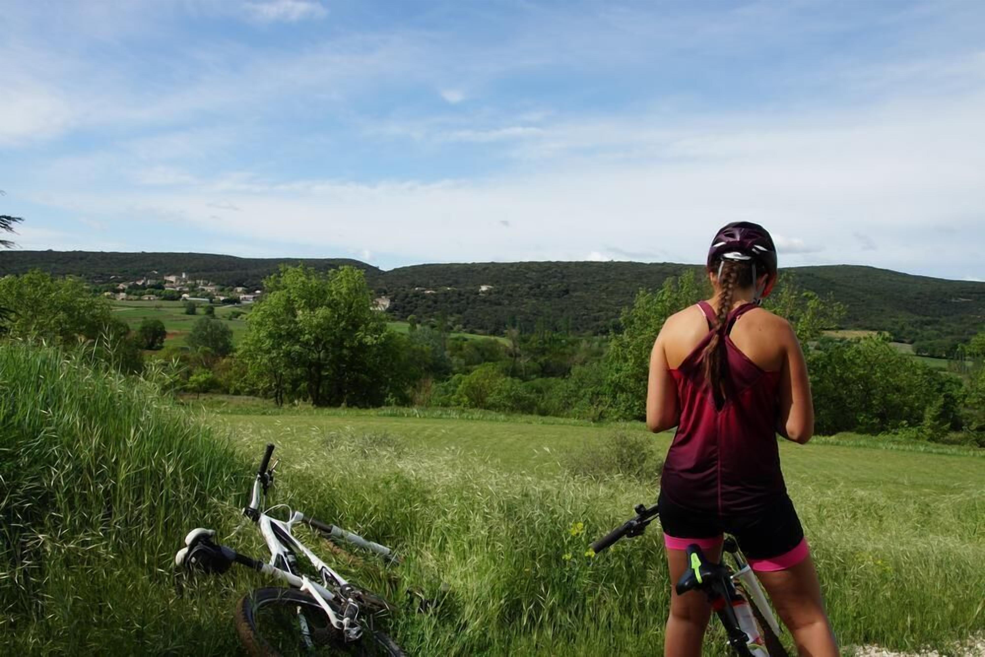 Una persona de pie en un campo de hierba, con una bicicleta, contemplando un paisaje montañoso con casas a la distancia.