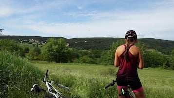 Une personne se tient dans un champ herbeux, à côté d’un vélo, et contemple un paysage vallonné avec des maisons qui se dessinent à l’horizon.