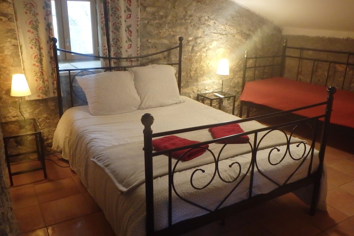 A bed with a metal frame, a red blanket, and a white bedspread in a room with stone walls and a window with floral curtains.