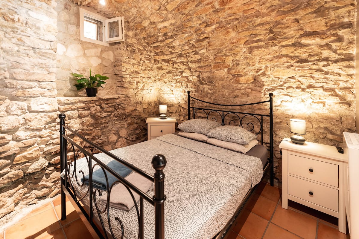 A bedroom with stone walls, a bed with a metal frame, two bedside tables with lamps, and a potted plant.