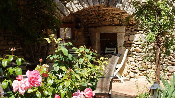 A stone building with a sign reading "Le Prieuré d'Orniols." There is a chair and a table outside, surrounded by greenery and rocks. - Prieuré d'orniols (Goudargues)