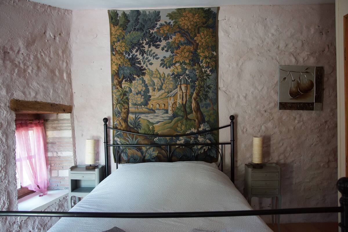 A bedroom with a bed, a tapestry, a nightstand, a lamp, and a window with curtains.