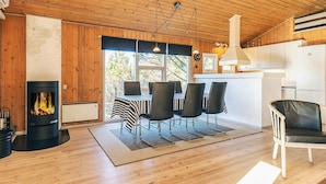 A cozy cabin interior with a wooden ceiling, a fireplace, a dining area with chairs, and a kitchen with white cabinets. - 4 Star Holiday Home in Albaek (Aalbaek)