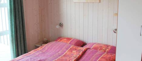 Una habitación con cama, lámpara de pared y ventana con cortinas.