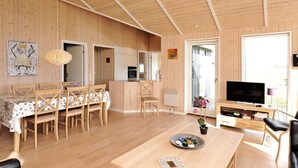 A spacious living area with wooden walls and ceiling, a dining table with chairs, a TV, and a kitchen area visible through an open door. - Modern Cottage With Spa - By Traum Ferienwohnungen (Sjølund)