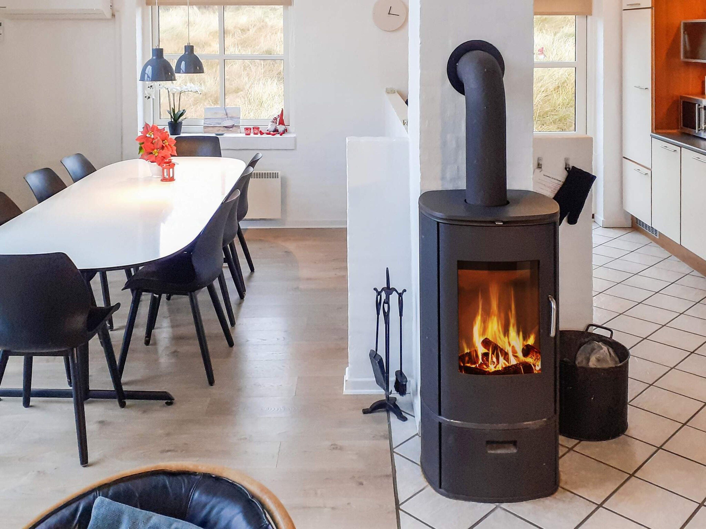 Une cuisine moderne avec un poêle à bois noir placé au centre, des meubles blancs et un coin repas comprenant une table blanche et des chaises noires.