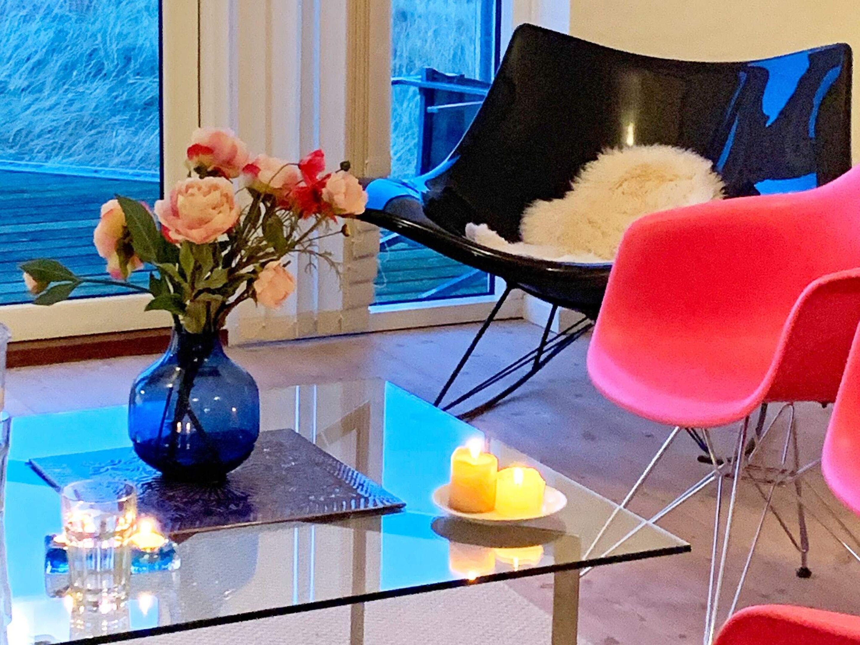 A modern room with a glass table, a vase with flowers, a chair with a fluffy pillow, and a red chair.