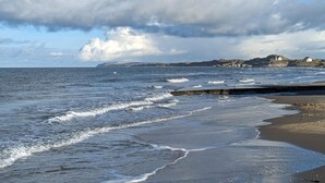 A coastal scene with waves, a sandy beach, and a distant rocky coastline with buildings.