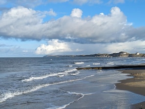 A coastal scene with waves, a sandy beach, and a distant rocky coastline with buildings. - 4 Star Holiday Home in Lokken (Lokken)