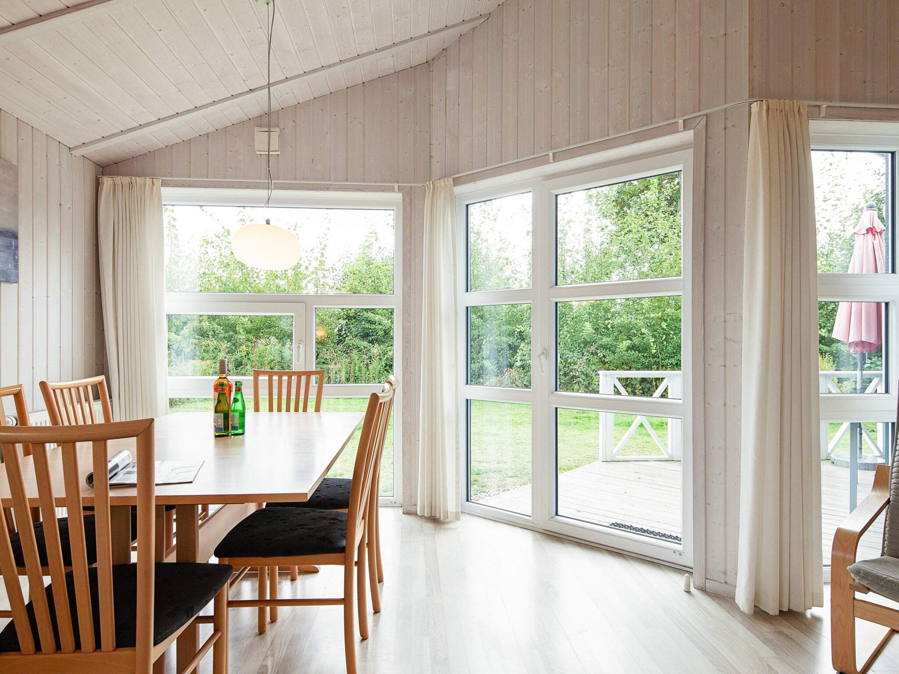 A dining area with a wooden table and chairs, large windows with curtains, and a view of greenery outside.