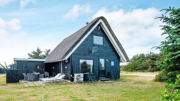 Eine dunkelblaue Holzhütte mit einem steilen Satteldach, einer Terrasse mit Gartenmöbeln und einem großen Fenster.