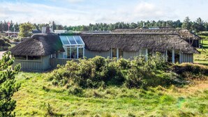 Un bâtiment traditionnel à toit de chaume, doté d’une extension en verre, et entouré de verdure.