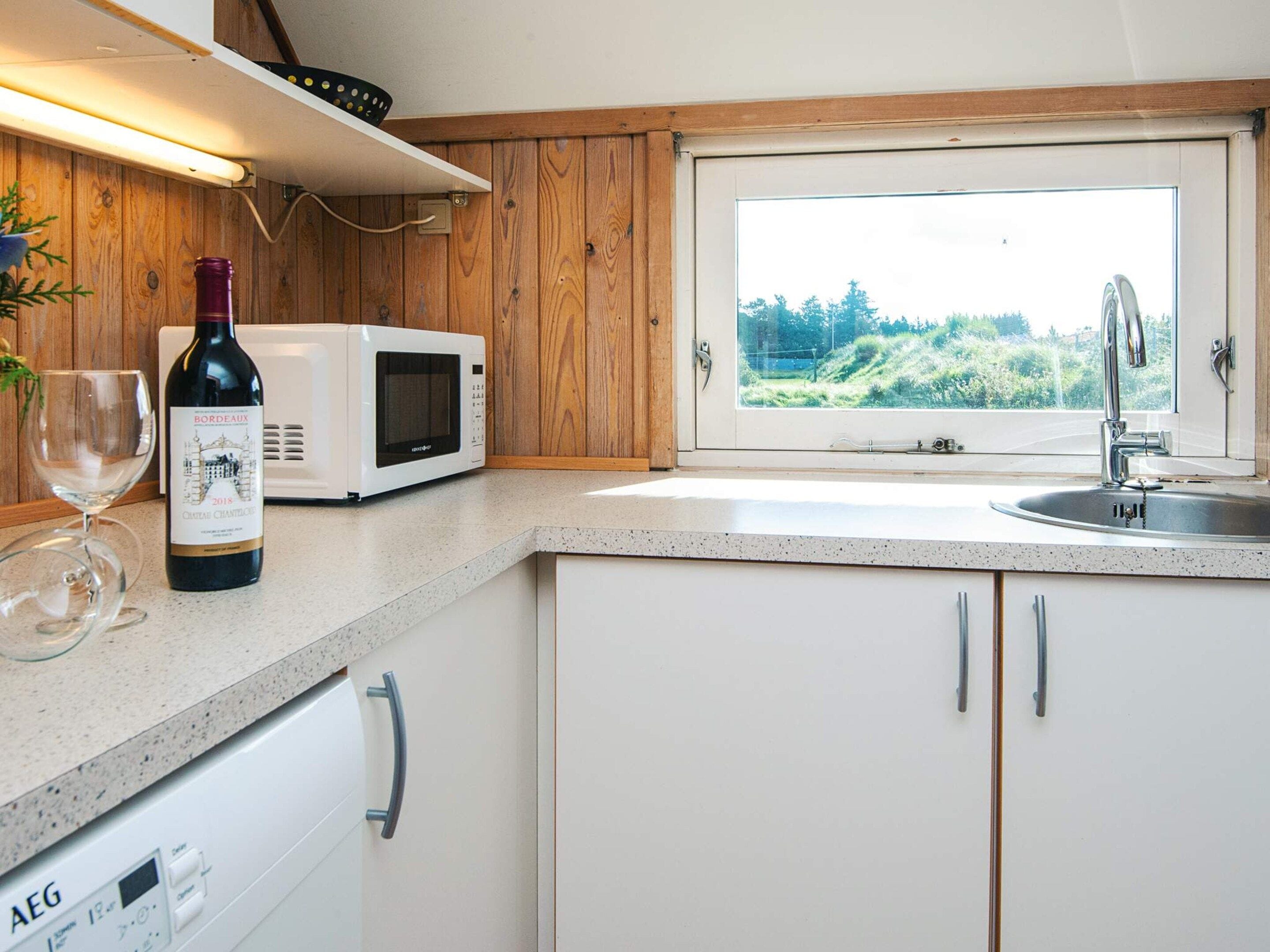 A kitchen with a microwave, wine bottle, and wine glass on the counter, a window with a view of greenery, and a dishwasher.