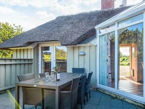 A patio with a table and chairs, a thatched roof, and a sliding glass door leading to a garden. - 4 Star Holiday Home in Vejers Strand (Vejers Strand)
