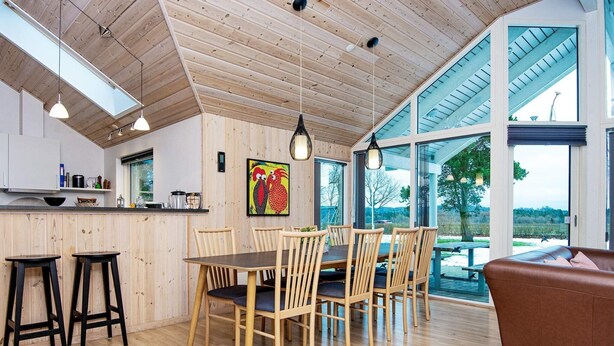 Eine moderne Küche mit Holzwänden, ein Essbereich mit Holztisch und -stühlen sowie große Fenster mit Blick ins Freie.