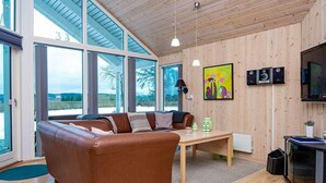 Ein modernes Wohnzimmer mit einem braunen Ledersofa, einem Holztisch und einem an der Wand montierten Fernseher. Große Fenster bieten einen Blick ins Freie.