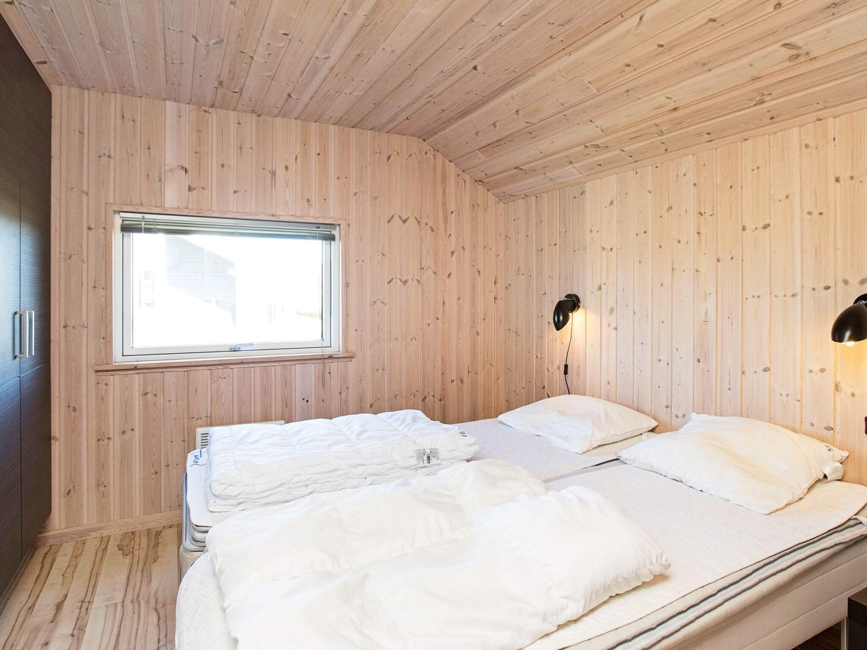A room with wooden panel walls, a window, two beds with white bedding, and a wall-mounted lamp.