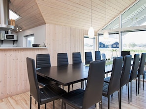 A modern dining area with a long table and black chairs, wooden walls, and large windows offering a view of the outdoors. - 5 Star Holiday Home in Idestrup (Idestrup)