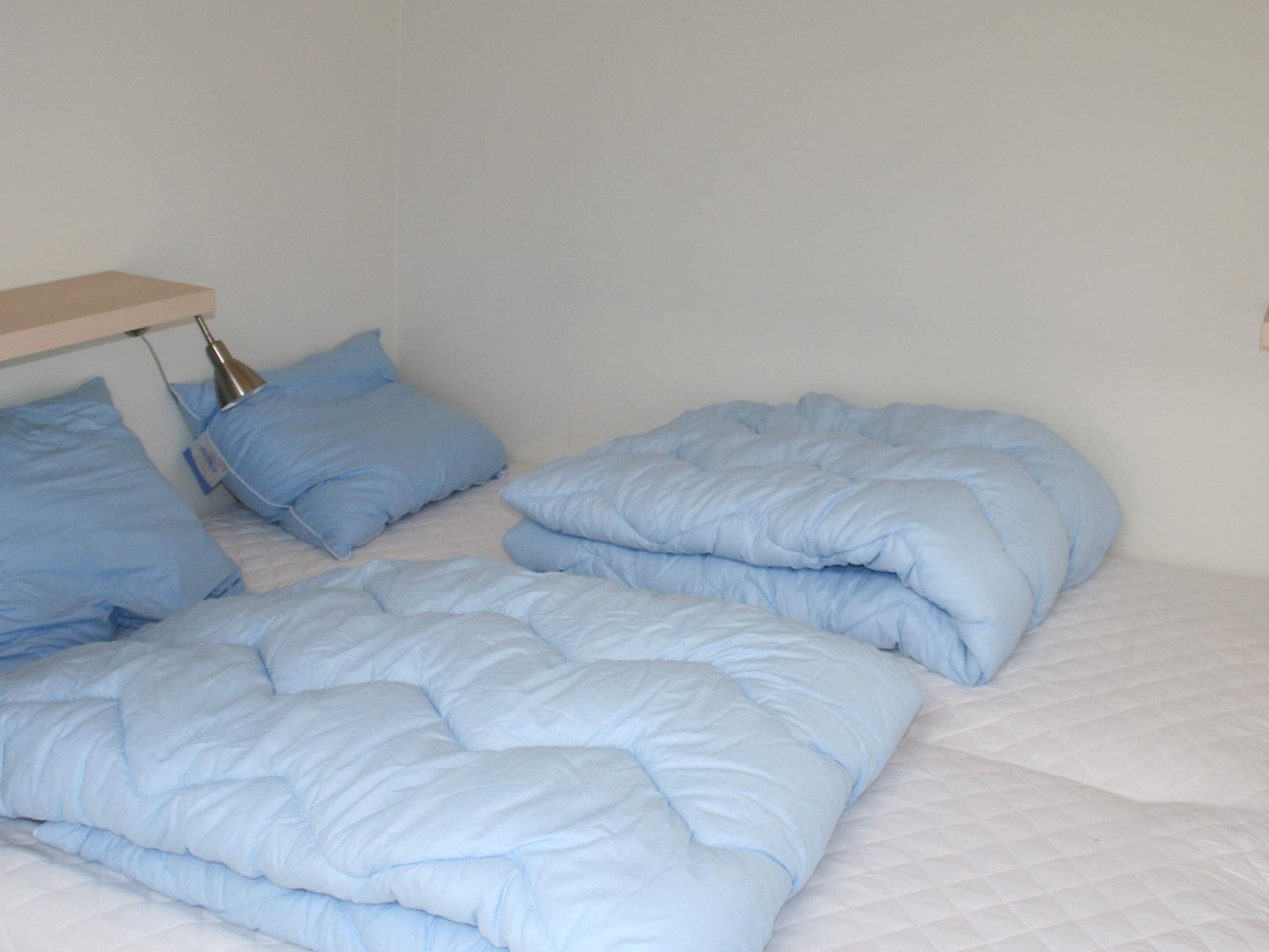 Ein Bett mit blauer Bettdecke und Kissen, ein Nachttisch mit Lampe und eine schlichte Wand.