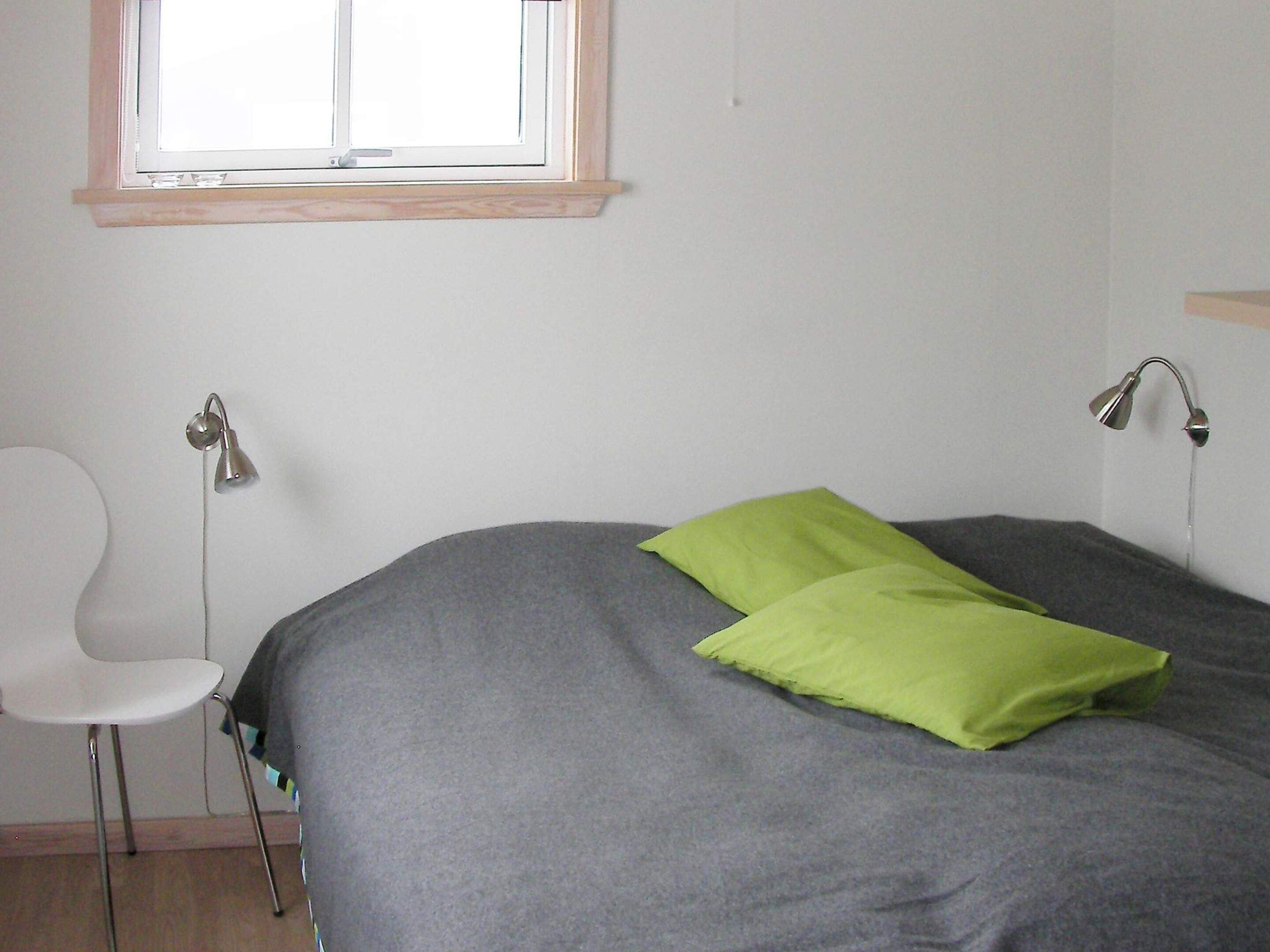 A room with a bed, a chair, a window, and a wall-mounted lamp.