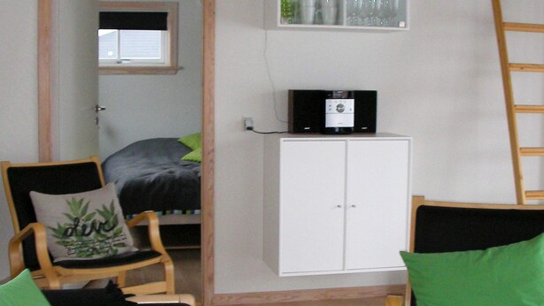 A room with a bed, a white cabinet, a chair with a green pillow, and a wooden ladder. - 6 Person Holiday Home in Gudhjem (Gudhjem)