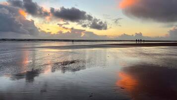 Une plage de sable humide qui reflète le ciel, quelques personnes qui se promènent et un coucher de soleil en arrière-plan.