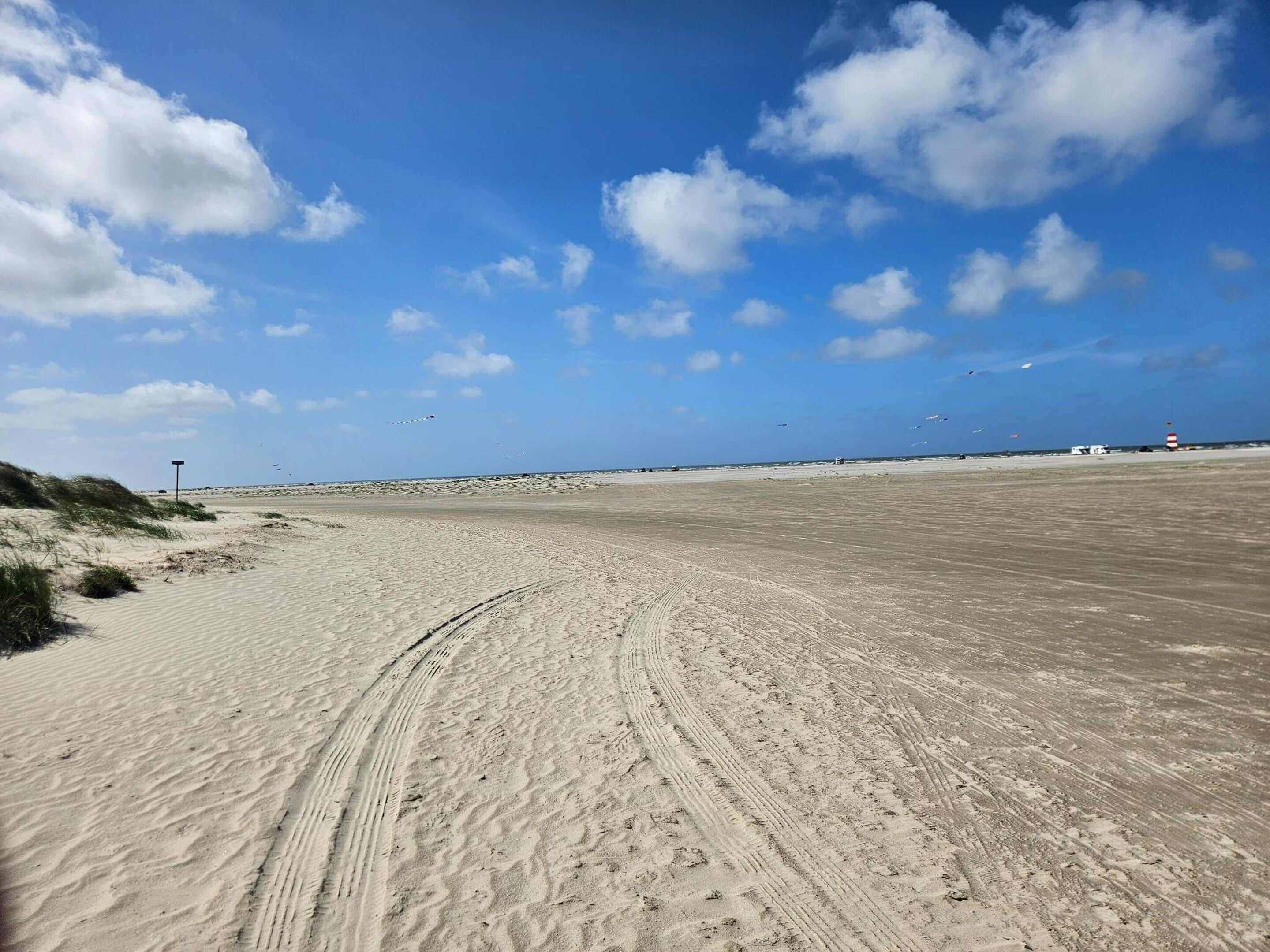 A wide sandy beach with tire tracks, a clear blue sky, and a distant lighthouse.