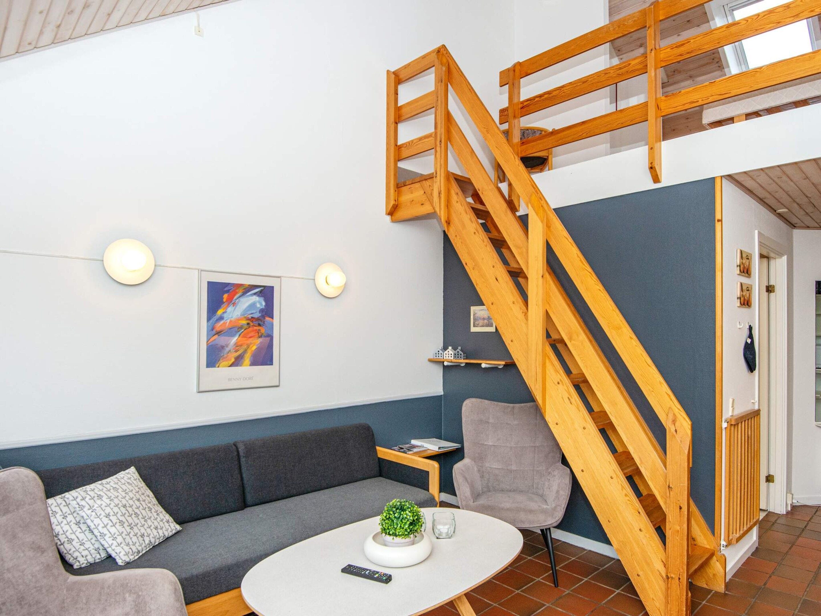 A modern living room with a grey sofa, a small table with a plant, a wooden staircase, and a framed artwork on the wall.