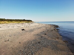 Uma praia com pedras e areia, céu claro e turbinas eólicas ao longe.