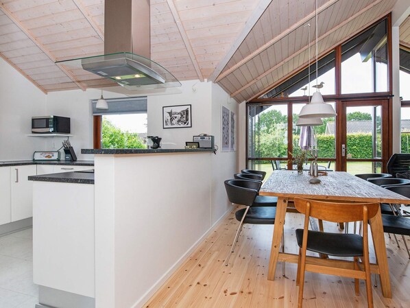 A modern kitchen with a central island, dining area with chairs, and large windows offering a view of greenery.