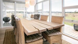 A modern outdoor dining area with a wooden table and wicker chairs, large windows offering a view of the landscape, and hanging pendant lights. - 6 Person Holiday Home in Brovst (Brovst)