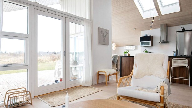 A spacious living room with a large sliding glass door leading to an outdoor deck, a cozy seating area with a white sofa and rattan chairs, and a kitchen area with modern appliances. - 6 Person Holiday Home in Brovst (Brovst)