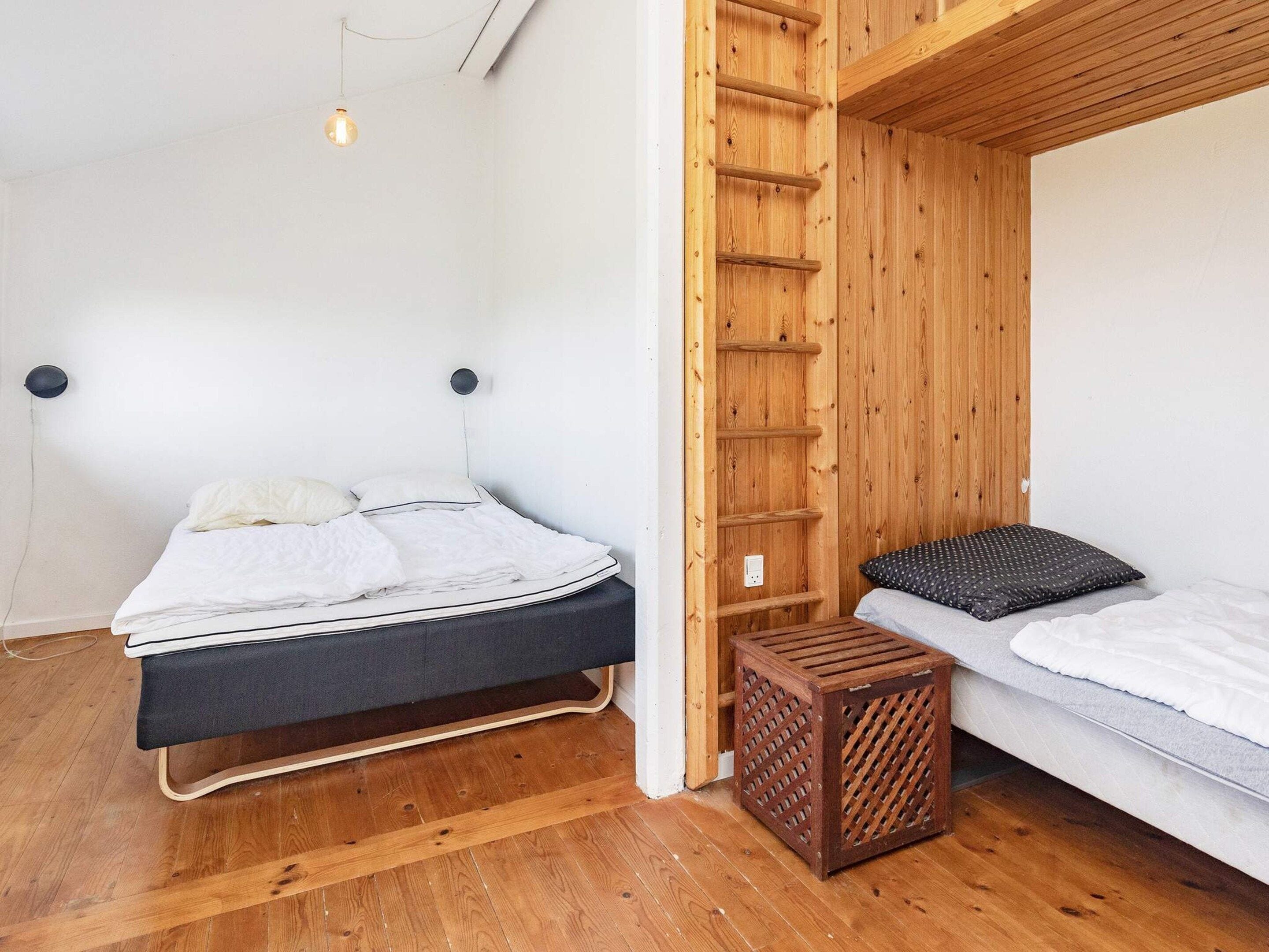 Quarto com duas camas, uma com estrutura preta e outra com cabeceira de madeira. Há um banquinho de madeira e uma prateleira fixada na parede.