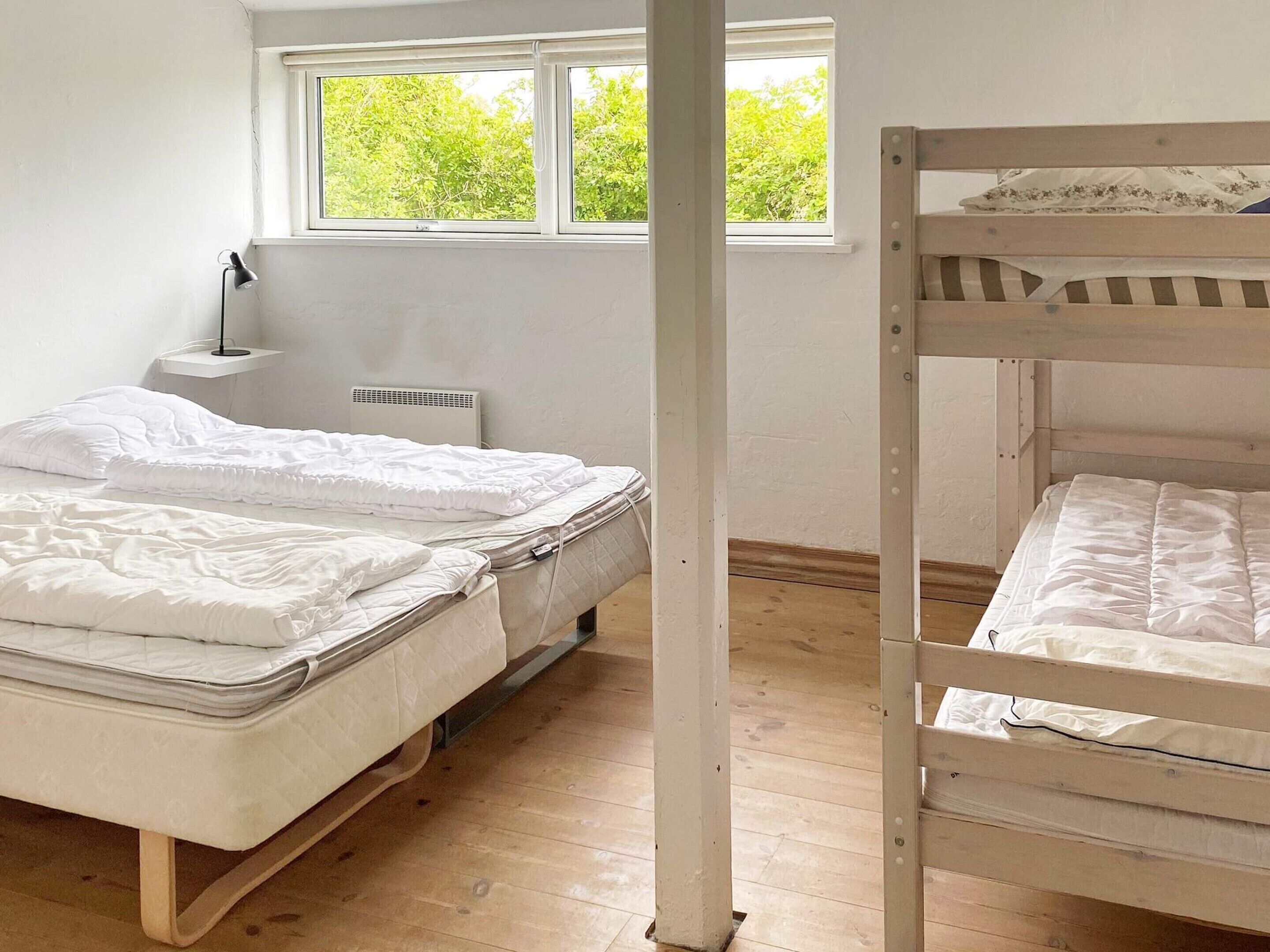 A room with a bunk bed, a single bed, a bedside table with a lamp, and a window with a view of greenery.