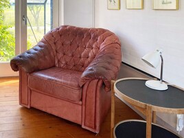 Un sillón de cuero marrón con respaldo acolchado, situado junto a una mesita redonda con una lámpara de escritorio blanca.