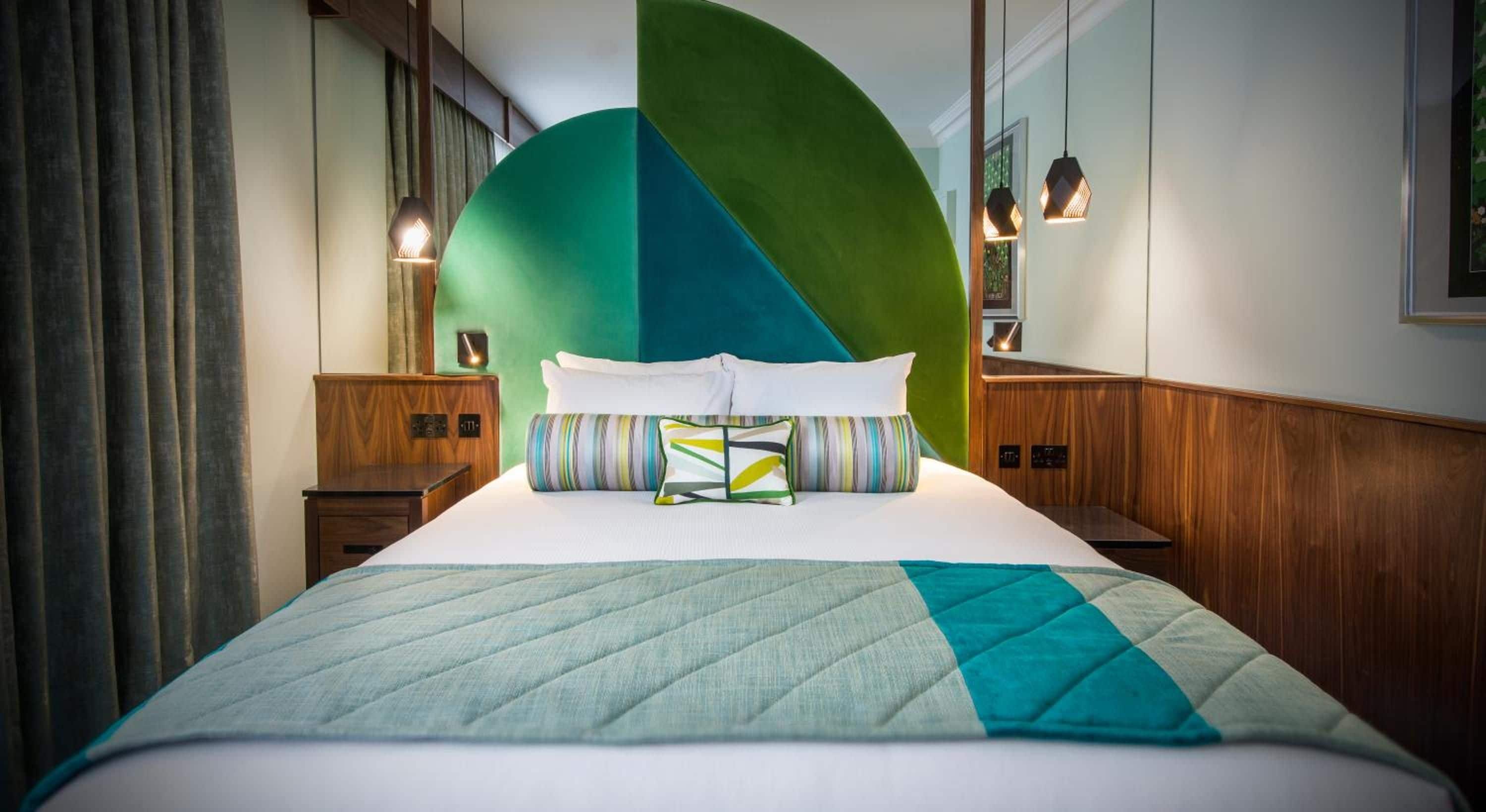 Habitación de hotel con una cama grande, pared verde y azul, paneles de madera y dos mesitas de noche con lámparas.