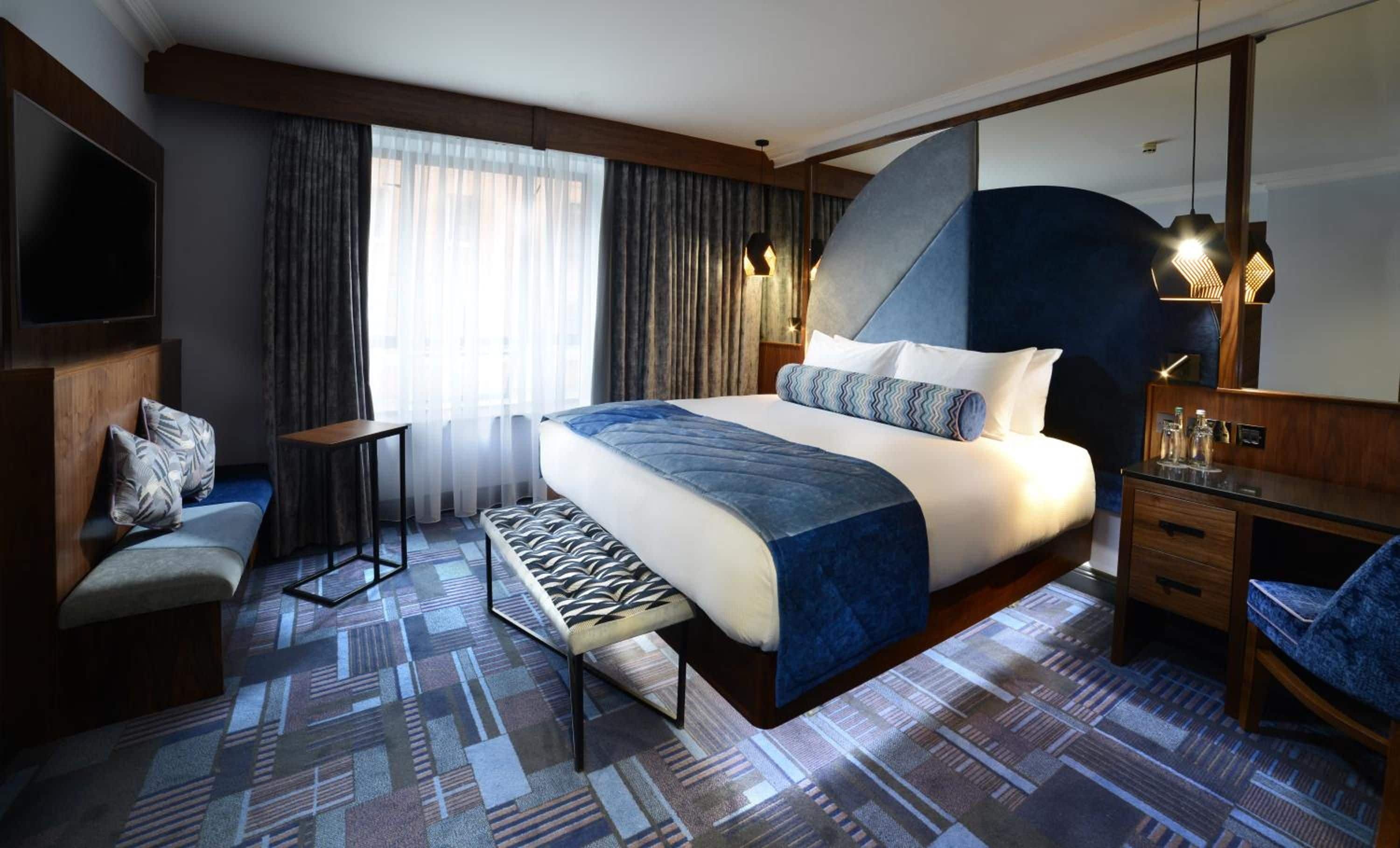 A hotel room with a large bed, a desk, a chair, a TV, and a window with curtains.