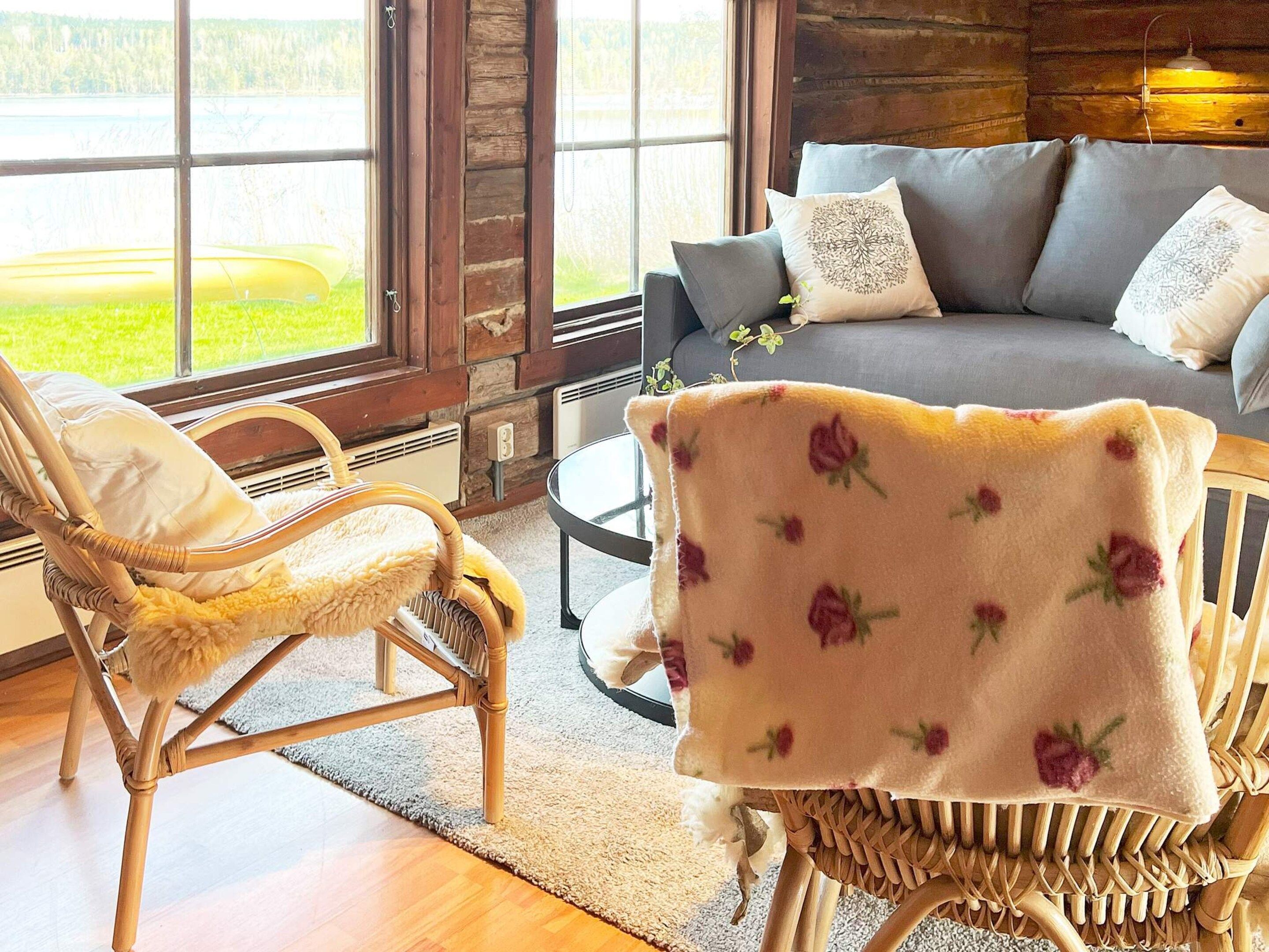 A cozy room with a wicker chair, a sofa with floral cushions, and a view of a green landscape through large windows.