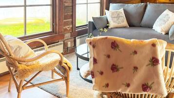 A cozy room with a wicker chair, a sofa with floral cushions, and a view of a green landscape through large windows.