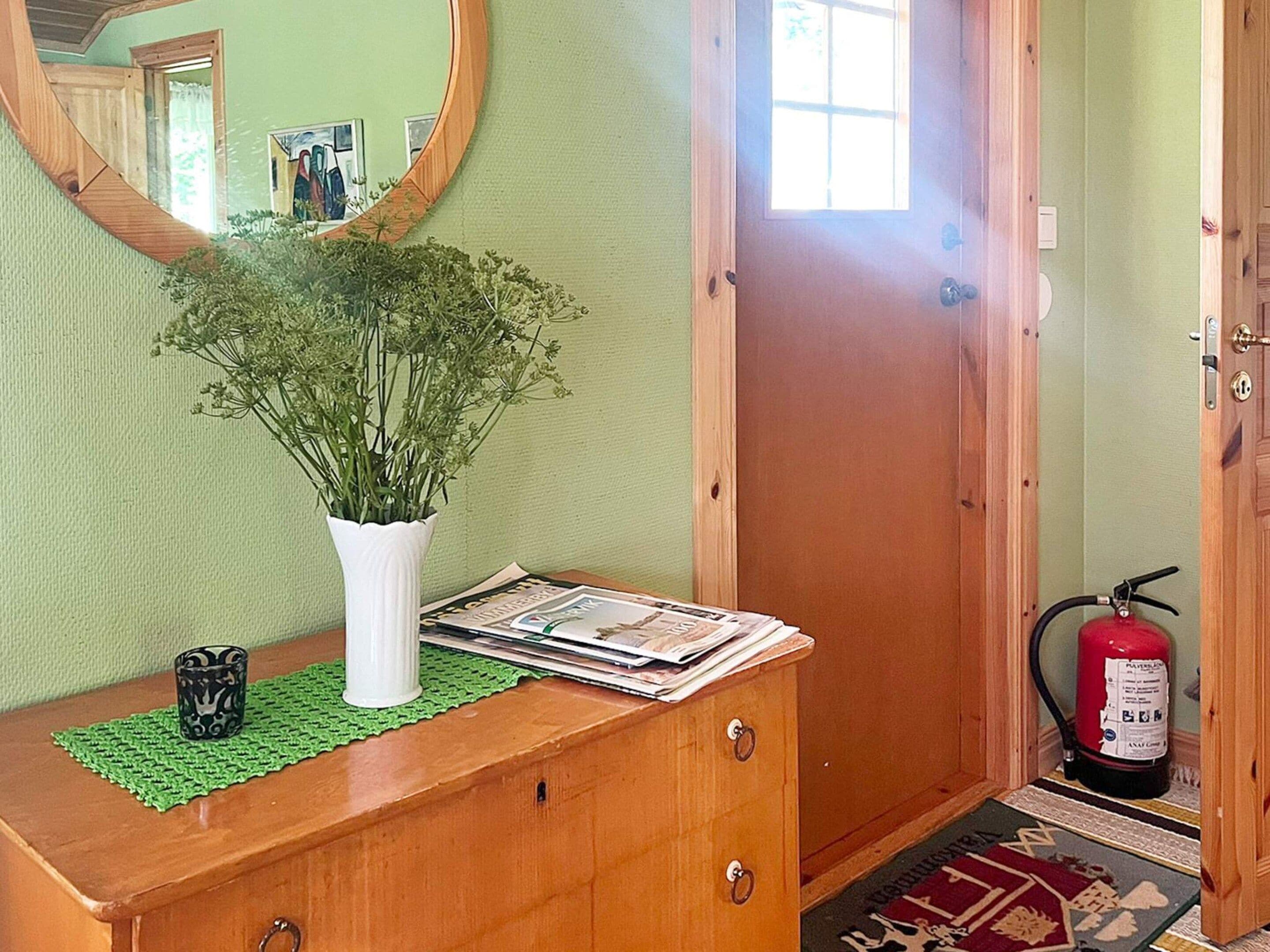 Un aparador de madera con un jarrón blanco lleno de plantas verdes, una revista y una taza negra. Un extintor está montado en la pared a la derecha.